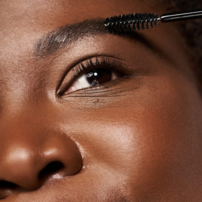 Young woman with radiant skin applies mascara, showcasing confidence and beauty in a studio.